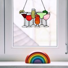 three martini glasses hanging from a window with a rainbow in the foreground and an orange, yellow, green, red, blue, pink, purple and white drink