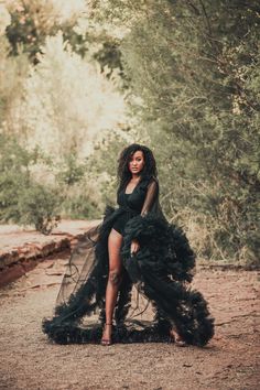 a woman in a black dress standing on a dirt road with trees and bushes behind her