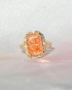 a ring with an orange stone surrounded by white and yellow diamonds on a pink background