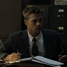 a man in a suit sitting at a desk writing on a piece of paper and holding a pen