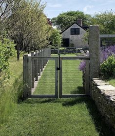 an open gate in front of a house with lavender flowers growing on the lawn and stone steps leading up to it