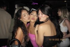 three beautiful young women standing next to each other at a party, one is kissing the other's cheek