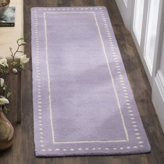 a purple rug with white trim on a wooden floor next to a vase and flowers