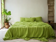 a bed with green sheets and pillows next to a potted plant on the floor