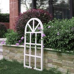 a white garden arch in front of a brick wall with purple flowers and bushes behind it