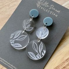 two clear earrings with leaves on them sitting on top of a gray card next to a pair of scissors