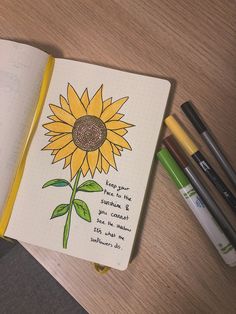 an open notebook with a drawing of a sunflower on it and some pens next to it