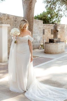 a woman in a wedding dress is walking down the street with flowers on her shoulder