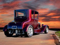 an old fashioned car driving down the road at sunset with clouds in the sky behind it