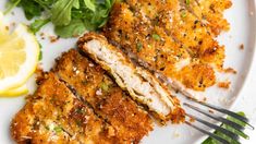 two pieces of chicken on a white plate with greens and lemon wedges next to it