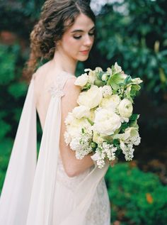 a woman in a wedding dress holding a bouquet of white roses and greenery with her back to the camera