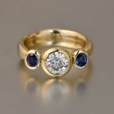 This is my contemporary interpretation of a classic 3 stone engagement ring. I treat the inside of the bezel settings with a rough sandblast which creates an added sparkle in the stones. This added feature makes the brightness comparable to that of a prong setting. I worked the shank in solid gold, subtly flaring out toward the bottom. This creates a flattering aesthetic. It also helps provide a counterweight to reduce the tendency of the ring to tilt around the finger. The focus is on using mor Saphire Jewelry, Engagement Ring With Sapphire, Jewelry Redesign, Stone Gold Ring, Sapphire Side Stones, 3 Stone Engagement Rings, Blue Bell, Right Hand Rings, Three Stone Engagement