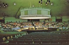 an auditorium filled with lots of people sitting at tables and standing in front of them