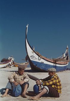 two men sitting on the beach next to boats