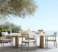 an outdoor dining table and chairs set up on a patio next to the ocean with olive trees in the background