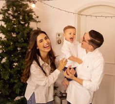 a man and woman holding a baby in front of a christmas tree with lights on it