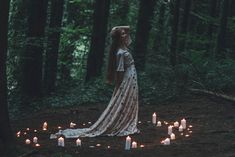 a woman standing in the middle of a forest with many candles around her and wearing a long white dress