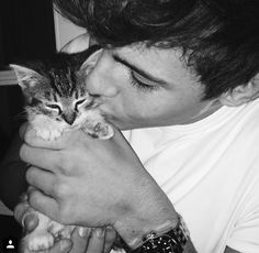 a man holding a kitten in his right hand and kissing it's face with the other hand