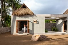 a woman standing in the doorway of a small house with thatched roof and walls