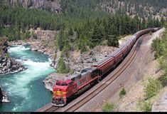 a red train traveling down tracks next to a river and forest covered mountain side area