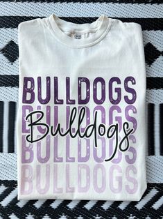 Get this popular new Bulldogs stacked text shirt and show off your school spirit in style!  Each shirt is printed in-house by me using high-quality materials on a Comfort Colors shirt, known for its exceptional comfort, style, and durability. Available in unisex sizes S to 3X, and youth sizes, we've got you covered regardless of your preferred fit. The Bulldogs stacked text Shirt features a classic crew neck and short sleeves, making it suitable for year-round wear. The flattering silhouette ensures a comfortable and stylish fit for both men and women. Please note that ivory is ONLY available in Adult sizes!  Note: As each shirt is individually printed, slight variations in color and placement may occur, adding to the uniqueness and charm of your Preppy Bulldogs Shirt. Your shirt will come Collegiate Style School T-shirt With Screen Print, Purple School Spirit T-shirt With Graphic Print, White Varsity Tops For School, School Spirit T-shirt With Graphic Print, White Varsity T-shirt For School, White Collegiate Tops For School, Casual Sublimation Design T-shirt With Letter Print For School, Collegiate Purple Tops With Letter Print, Varsity Style Letter Print T-shirt For School