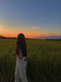 a woman standing in the middle of a field at sunset with her back to the camera