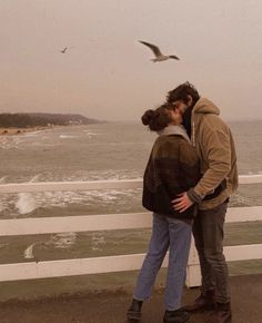 a man and woman standing next to each other on a pier near the ocean with seagulls flying overhead