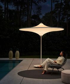 a man sitting in a chair under an umbrella next to a swimming pool at night