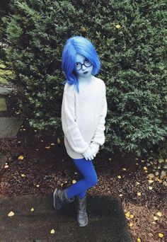 a person with blue hair and glasses standing in front of a bush