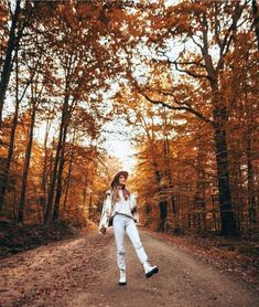 a woman standing in the middle of a dirt road surrounded by trees with yellow leaves