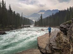 a woman standing on the edge of a cliff next to a river and forest area