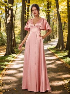 a woman in a long pink dress stands on a path surrounded by trees and leaves