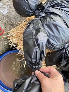 a hand is holding a black trash bag over a blue bowl and some straw on the ground