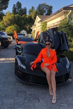 a woman in an orange dress sitting on top of a black sports car with the hood open