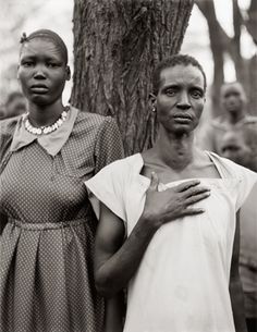 two women standing next to each other in front of a tree and people looking at the camera