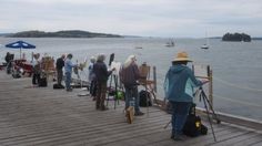 several people standing on a pier near the water with umbrellas and other items in front of them