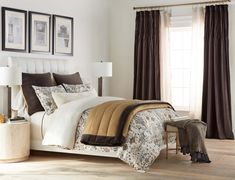 a bedroom with white walls and brown drapes on the windowsills, a large bed in front of two framed pictures