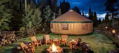 a yurt in the woods lit up at night with chairs around it and fire pit