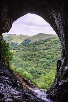 the view from inside a cave looking down at trees and hills in the distance with a river running through it