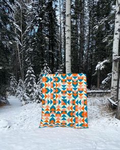 an orange and blue quilt sitting on top of snow covered ground in front of trees