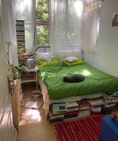 a bed with green sheets and pillows in a small room next to a window filled with books