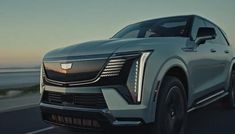 the front end of a silver cadillac suv driving on a road near the ocean at sunset