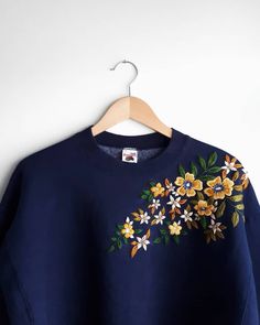 a blue sweatshirt with yellow and white flowers on the front, hanging from a wooden hanger