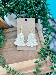 the white christmas tree earrings are sitting on top of a wooden board next to some green leaves