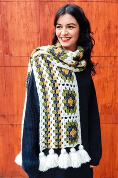 a woman standing in front of a wooden wall wearing a crocheted scarf with tassels