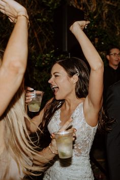 two women in white dresses are holding drinks and dancing with their arms above their heads