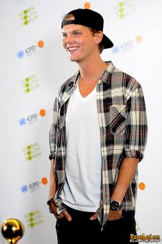 a young man standing in front of a white wall wearing a plaid shirt and black hat