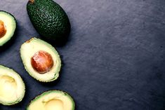 an avocado cut in half and whole on a black surface with other fruits