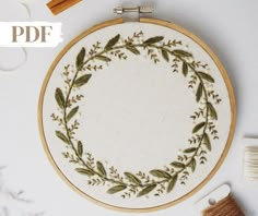 an embroidered wreath with green leaves and sprigs sits on a white surface next to some thread