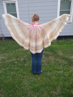 a woman is standing in the grass holding up a crocheted shawl